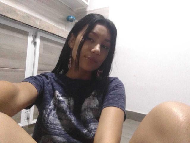 KataleyaMansson with dark hair, nose piercing, wearing a blue graphic shirt, sitting with legs up, smiling slightly, wearing long earrings, in a room with wooden panels.