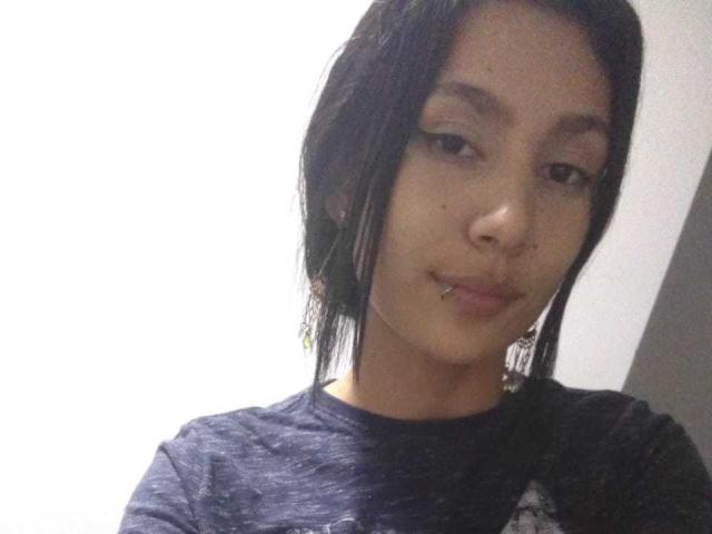 KataleyaMansson with straight black hair, subtle makeup, and a small lip piercing, wearing a dark blue shirt, smiling slightly, looking at the camera.