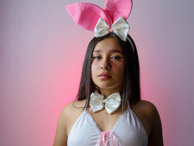 LilithSwett with pink bunny ears, white bowtie, and white halter top, showing small tits, long brown hair, and a nose piercing, under pink and white light.