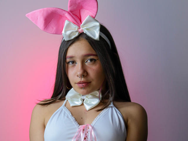 LilithSwett with pink bunny ears, white bow, and choker, wearing a white halter top, has a nose ring and lip piercing, looking cute and sexy.