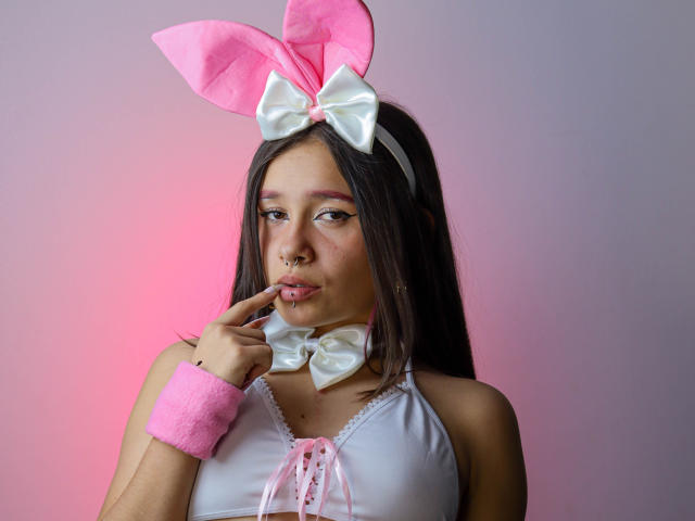 LilithSwett with pink bunny ears, white bow, and pink wristband, biting finger, nose ring, pink lipstick, white top, long brown hair, pink and white gradient background.