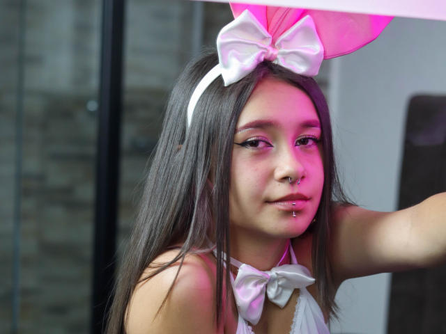 LilithSwett with long brown hair, white bow headband, white bow tie, nose ring, and pink-lit face, showing off her cute, sexy, small tits, and pierced lip.