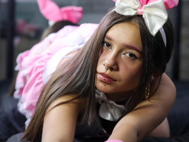 LilithSwett, with long brown hair, wearing a pink and white bunny costume, has a nose ring, lip ring, and a gold earring, lying on her stomach, looking at the camera.