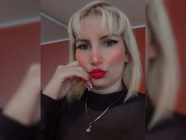 Giulia Addams bionda, con labbra rosse, una maglia nera trasparente, una collana dorata e unghie dipinte, che tocca la guancia, riflessa in uno specchio. Il suo seno è parzialmente visibile attraverso la maglia..