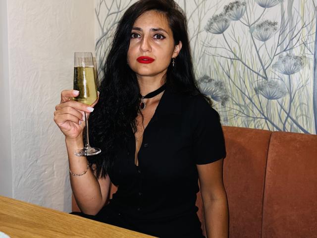 JennaEasy with long black hair, red lipstick, black dress, holding champagne glass, sitting in brown booth, wooden table in front, looking serious, wearing black choker, bracelet on right wrist.