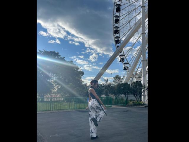 SalomeMille stands with her back to the camera, wearing a white floral dress, holding a stick, and a sunbeam shines through her brown hair. She's near a Ferris wheel.