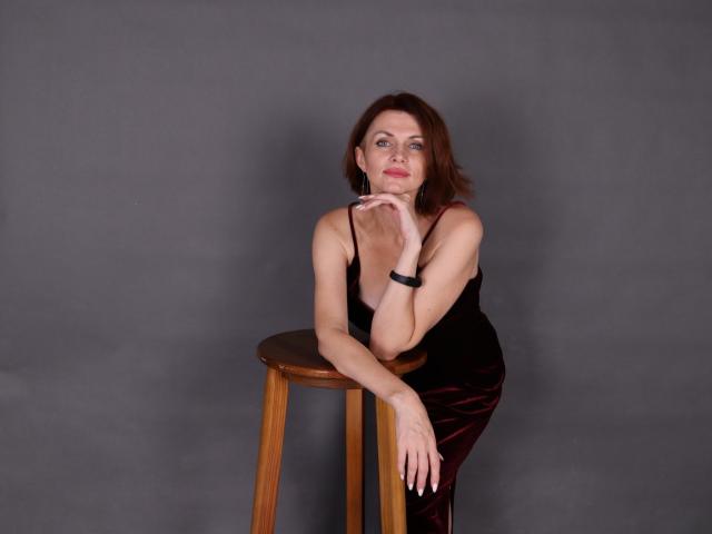 LisaTreds with red hair, black velvet dress, sitting on wooden stool, hands on her chin, gray background, looking at camera, wearing black bracelet, medium tits, slim build.