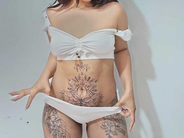 Salma, con un bikini blanco con costillas, mostrando tatuajes: un tatuaje floral grande en el abdomen, tatuajes florales en los muslos. Manos tirando hacia abajo de la parte inferior del bikini. Pechos desnudos, cabello castaño..
