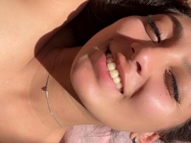DianaFernandezz's face, sunlit with closed eyes, smiling, showing her teeth, with a thin necklace, and her brown hair spread out. Her ass is partially visible.