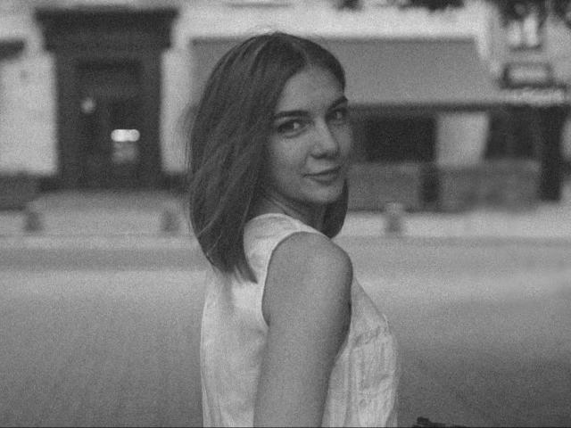 Black-and-white photo of LoveisSense-hot with shoulder-length hair, looking over her shoulder, wearing a sleeveless top, standing on a street, showing her small, perky ass.