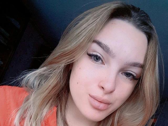 LoveisSense-hot with blonde hair, light skin, and full lips, wearing an orange shirt, has thick eyebrows, long eyelashes, and a small mole on her right cheek, looking at camera.
