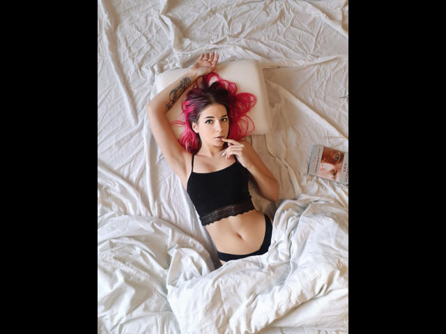 LoveisSense-hot with pink hair, black lace top, and black panties, lying on white sheets, holding a cigarette, arm tattoo visible, book to the right.