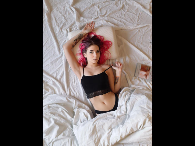 LoveisSense-hot with pink hair, black lace-trimmed top, and black panties, lying on a white bed, arms above head, looking at camera, tattoos visible, book to the right.