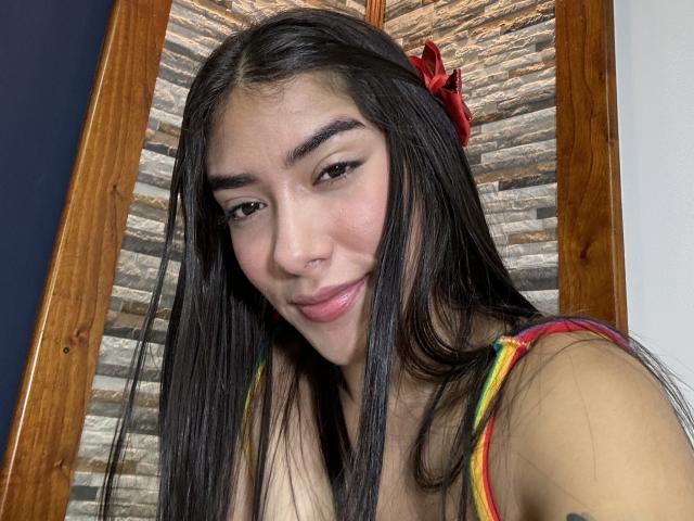 SharonKlart with long black hair, red hair clip, and a subtle smile, wearing a rainbow tank top, showing her small tits and cute face. Her skin is tan.