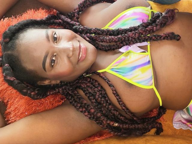 DannaJonez with dark brown skin, red-brown braided hair, smiling, wearing a colorful rainbow bikini, lying on an orange pillow, showing her tits.