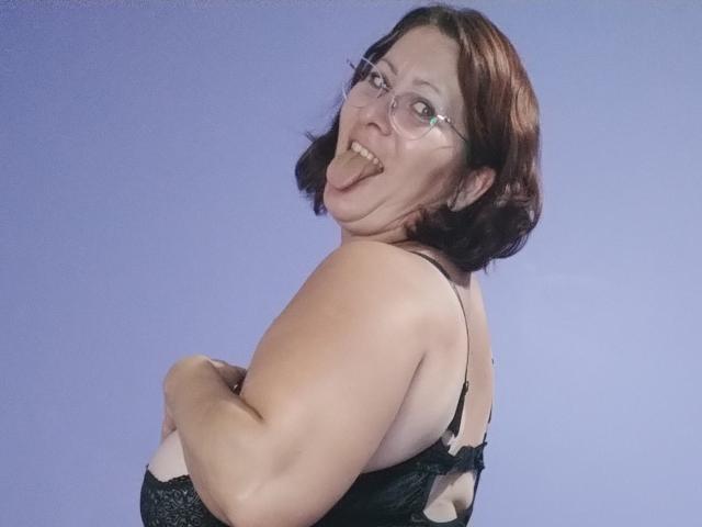 SoniaLaBelle with short brown hair, glasses, and black lace bra, sticking out her tongue, arms crossed, showing her big ass and small tits against a blue background.