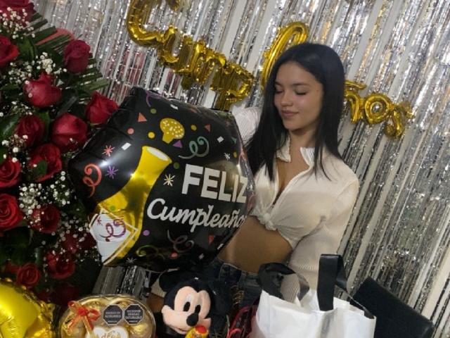 IssaPuasini with black hair, white shirt, holding black "Feliz Cumpleaños" balloon, surrounded by red roses, Mickey Mouse, gold balloons, and silver streamers.