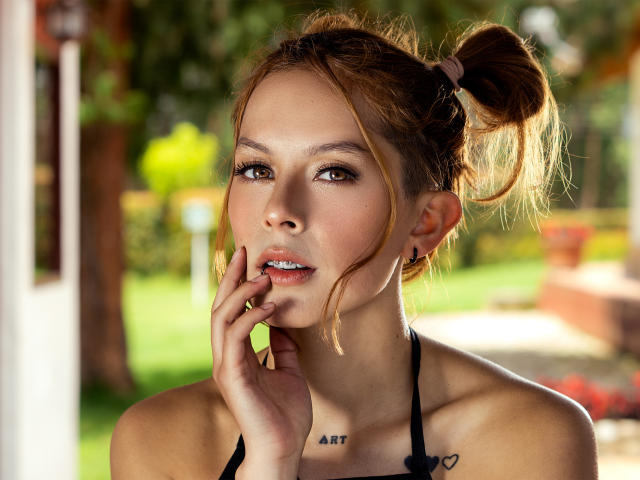 VictoriaOrtega has light brown hair in a messy ponytail, brown eyes, and a small tattoo under her chin. She's touching her lip with her finger, wearing a black halter.