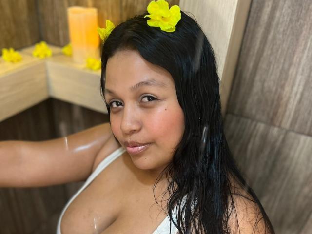 EmilyTerrys with wet black hair, yellow flowers in her hair, white tank top, showing her big boobs, smiling slightly, in a wooden tiled bathroom, candles in the background.