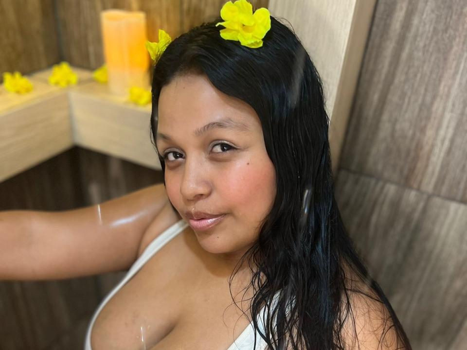 EmilyTerrys with wet black hair, yellow flowers in her hair, white tank top, showing her big boobs, smiling slightly, in a wooden tiled bathroom, candles in the background.