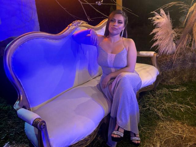 BellaSmithX69 sitting on a vintage blue-and-white sofa, wearing a grey, lace-trimmed dress, holding a purple drink, with her tits slightly visible, and her ass resting on the seat.
