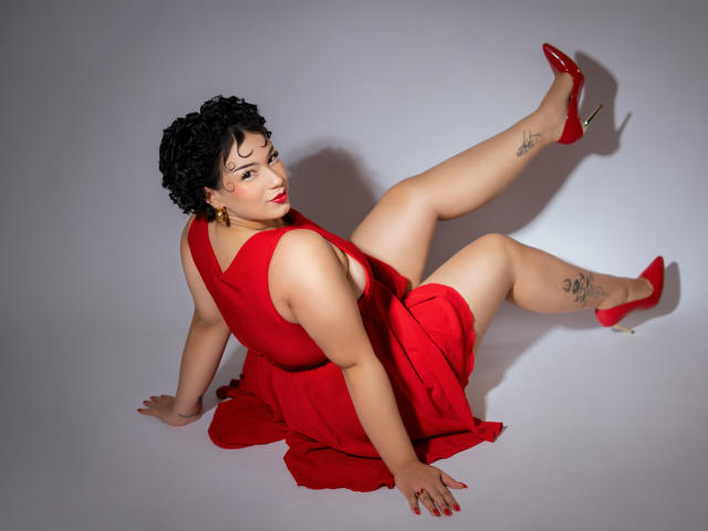 Milly Johnson, volslank, met kort zwart krullend haar, draagt een rode jurk en rode hakken, heeft tatoeages op haar dij, zit met één been opgetrokken en laat haar achterwerk zien..