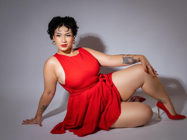 Milly Johnson, de figura voluptuosa, con cabello negro rizado corto, vestido rojo, que muestra sus grandes senos y sus redondeadas caderas, tatuajes en el brazo y el muslo, tacones rojos, sentada en el suelo blanco..