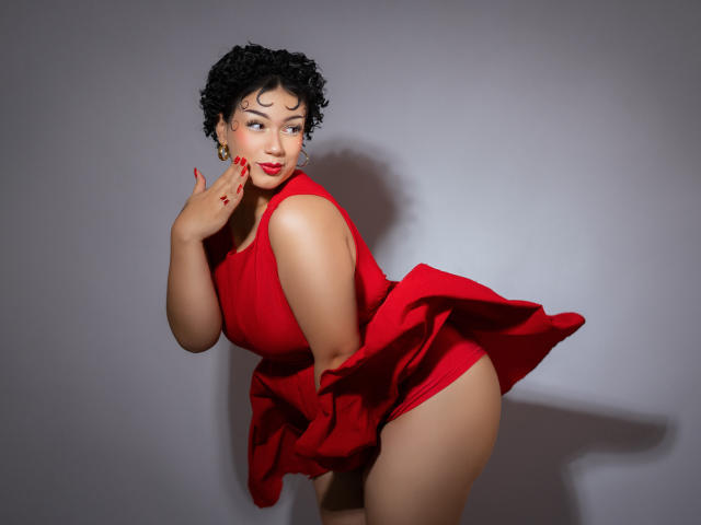 Milly Johnson, com cabelo preto encaracolado, vestido vermelho e batom vermelho, inclinada para frente, mostrando o bumbum, sorrindo com a mão perto do rosto, unhas vermelhas, fundo cinza, iluminação de estúdio..