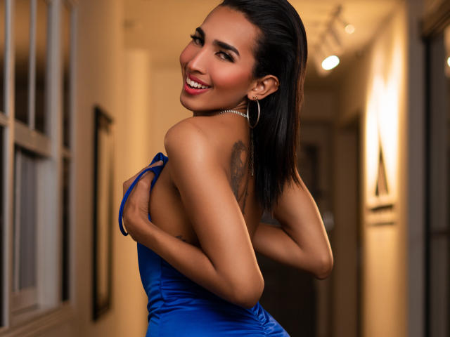 GabySantos with black hair, smiling, wearing a blue dress, holding her tits, showing a tattoo on her back, in a hallway, wearing hoop earrings, looking over her shoulder.