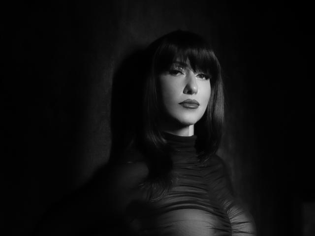 Foto em preto e branco de MissVonTease, com cabelo preto e liso, franja e expressão séria, vestindo uma blusa transparente com babados, que realça sutilmente seus seios. Fundo escuro..