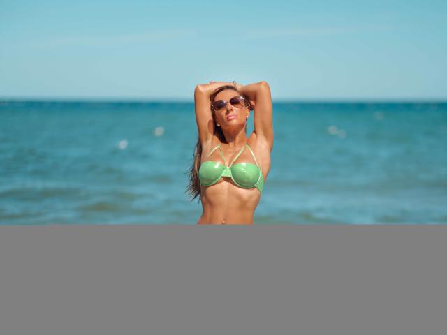 JoannaBraun in a green bikini, standing in shallow water with arms raised, showing her tits and ass, wearing sunglasses, with a tan, curvy body and long brown hair.