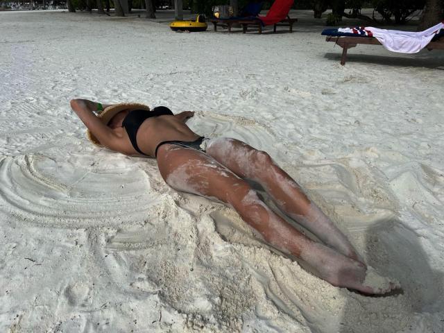 AliceMarr lies face-down on white sand, wearing a black bikini with her ass and legs covered in sand, arms above her head, sunbathing on a beach.