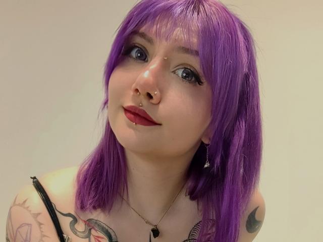 PerfectGabbie, con cabello morado, aro en la nariz, piercing en el tabique, lápiz labial rojo y tatuajes en los hombros, mirando hacia arriba con ojos azules, vistiendo una camiseta negra con tirantes..