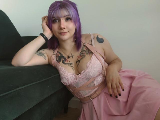 PerfectGabbie met paars haar, zwarte nagellak, een roze kant beha en een roze jurk, die haar tatoeages en zwarte horloge laat zien, leunt tegen een groene bank en glimlacht..