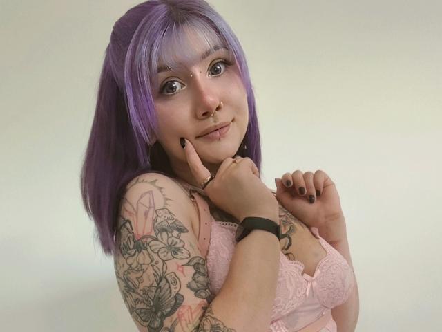 PerfectGabbie, con cabello morado, aro en la nariz y tatuajes, sosteniendo su barbilla, vistiendo un sujetador de encaje rosa, una pulsera negra y esmalte de uñas oscuro, sonriendo suavemente..