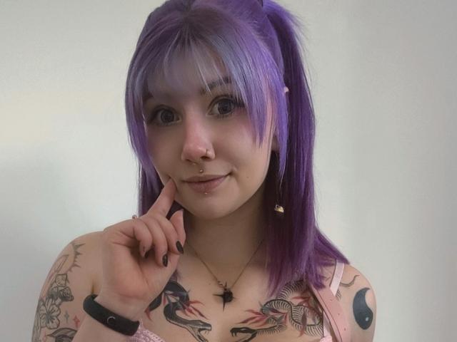 PerfectGabbie with purple hair in pigtails, nose ring, and lip piercing, touching her chin, showing tattoos on her arms and chest, wearing a pink top, black wristband.