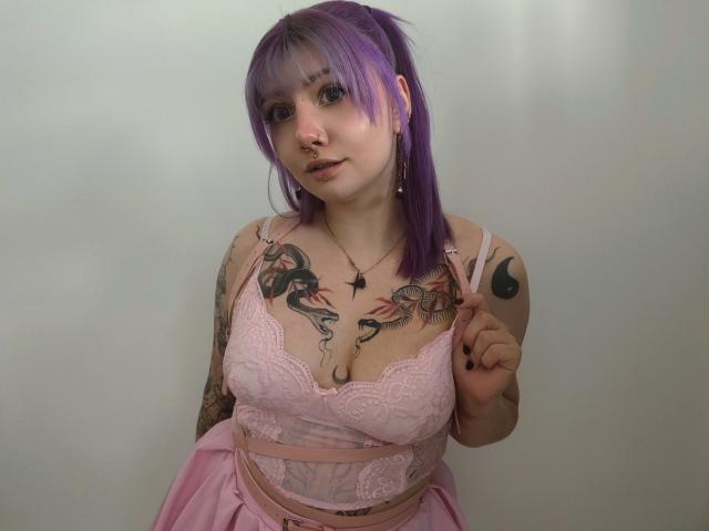 PerfectGabbie with purple hair, nose ring, and tattoos, wearing pink lace bra and skirt, showing tits, making a peace sign, holding straps, and looking at camera.