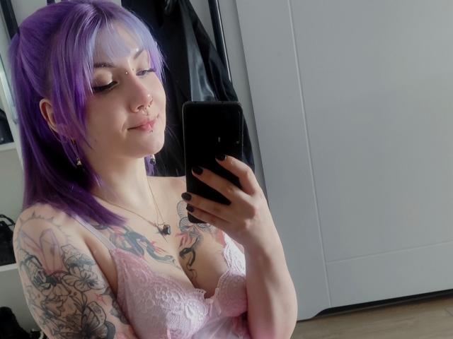 PerfectGabbie met paars haar, neusring en tatoeages, die een selfie maakt in een spiegel, gekleed in roze kant lingerie, waarbij haar borsten zichtbaar zijn, en een zwarte telefoon vasthoudt..