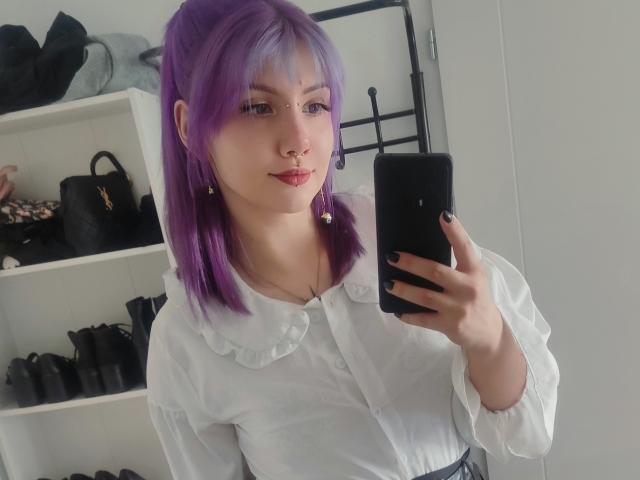 PerfectGabbie met paars haar, een neusring en oorbellen, die een witte blouse draagt, een telefoon vasthoudt, een spiegel-selfie maakt, haar volle lippen laat zien en zwarte nagellak heeft..