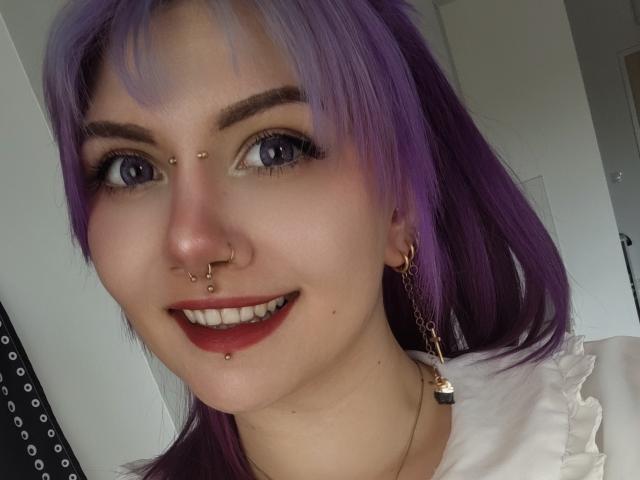 PerfectGabbie with purple hair, blue eyes, nose piercings, lip piercing, red lipstick, smiling, white shirt, gold earrings, and a chain with a small pendant.
