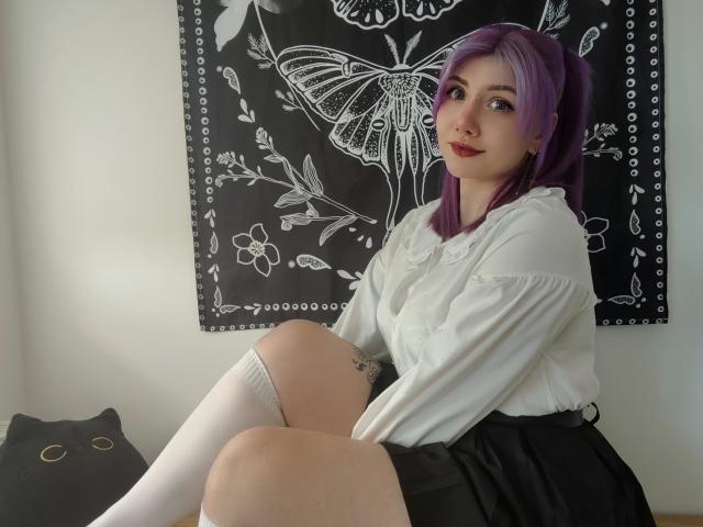 PerfectGabbie met paars haar, een witte blouse, een zwarte rok, witte kniekousen, zittend met de knieën opgetrokken, met een tatoeage zichtbaar op haar linker dij, en een schattig zwart kattenkussen..