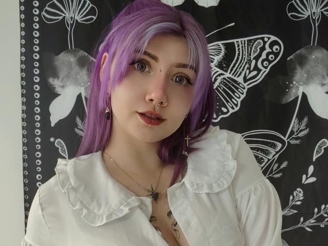 PerfectGabbie with purple hair, nose ring, and cross earrings, wearing a white ruffled shirt, has puffy lips and a tattoo under her chin, looking at the camera.