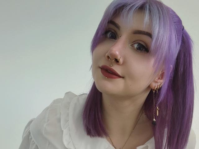 PerfectGabbie has purple hair, a nose ring, and lip piercing, wearing a white shirt with ruffled sleeves, looking at the camera with a slight smile.