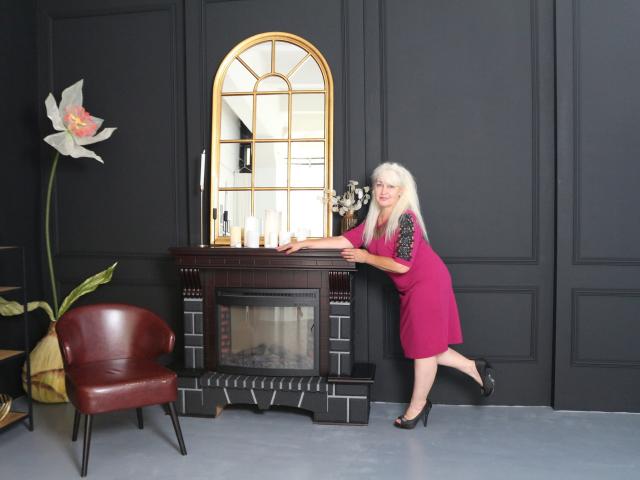 KamaliaSim with long blonde hair, pink dress, and black heels, leans on a fireplace, showing off her tits, with a red chair and large white flower to the left.