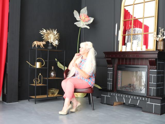 KamaliaSim with long blonde hair, plaid shirt, pink pants, and beige heels, sitting on a chair, holding a large flower, in front of a dark fireplace.