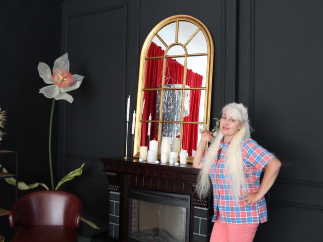 KamaliaSim with long white hair, wearing a pink and blue checkered shirt, standing by a black fireplace with candles, and a gold-framed mirror reflecting red curtains.