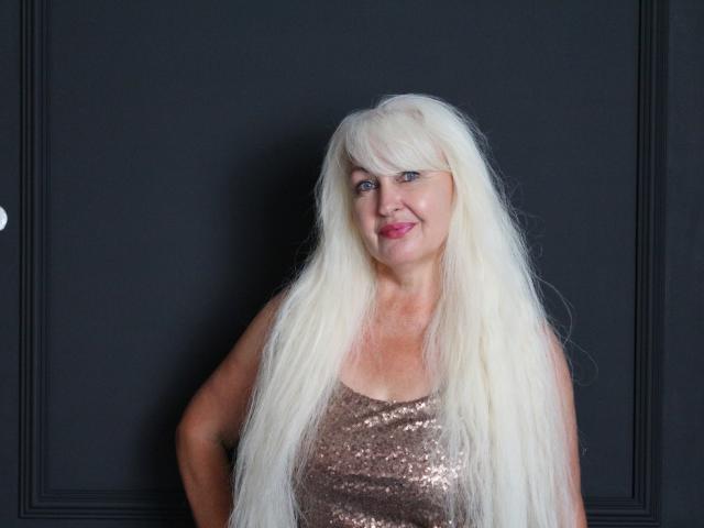 KamaliaSim with long, white, wigged hair, wearing a shiny, pink, sequined tank top, standing against a dark background, smiling with pink lipstick. Boobs visible under top.