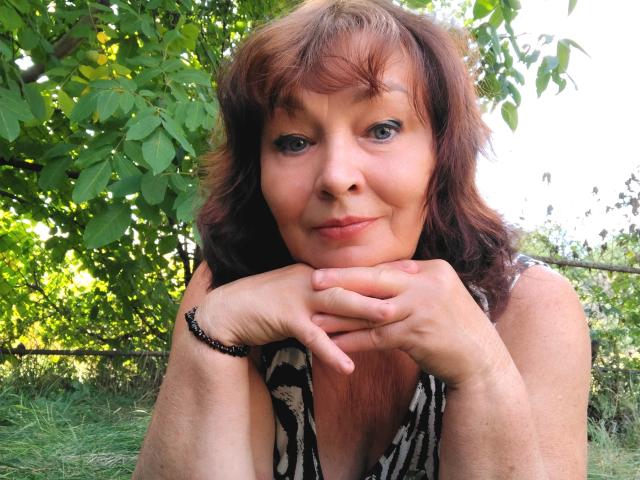 MilaMils with wavy brown hair, blue eyes, and a slight smile, resting her chin on her hands, wearing a zebra-patterned top, and a black bracelet, outdoors.
