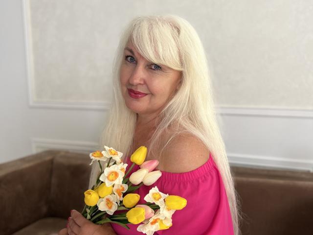 KamaliaSim with long blonde hair, pink off-shoulder top, holding yellow and white tulips, smiling with red lipstick, sitting on brown sofa, white wall in background.