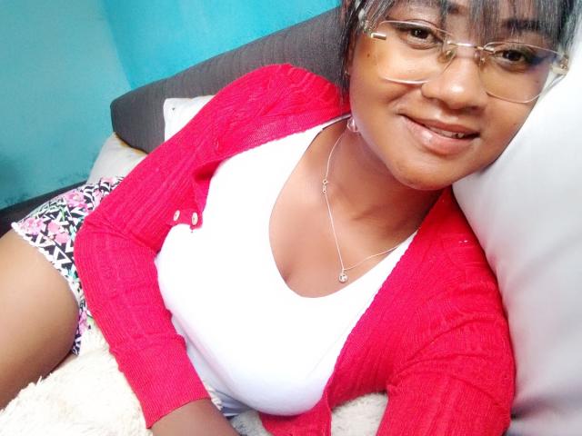 MelissahHot with medium brown skin, glasses, and a smile, wearing a red cardigan, white tank top, and floral shorts, lying on a white pillow. Her tits are small.